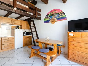 Eine Küche mit Holzschränken, einem weißen Kühlschrank, einer Mikrowelle und einem Herd. Ein Holzessttisch mit blauen Kissen und ein Fernseher auf einer Holzkommode.