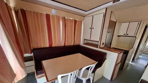 A compact kitchenette with a small table and chairs, a dark-colored bench, and a window with curtains. - Boat stay by the Mayenne - 9 guests (Chenillé-Changé)