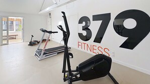 Een ruime fitnessruimte met een loopband, elliptische trainer en een hometrainer, en een groot bord met het nummer 379.