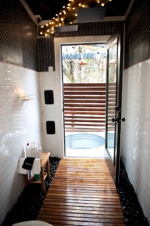 Un baño con piso de madera, paredes de azulejos blancos, un banco y una puerta de vidrio que da a un área exterior con piscina.