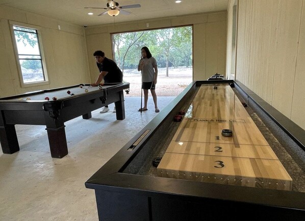 Une table de billard et une table de jeu de palet sont installées dans une pièce dotée de grandes fenêtres et d’un ventilateur de plafond.