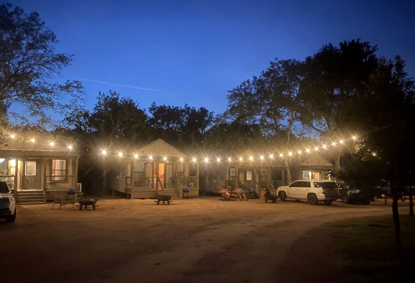 A rustic cabin-style building with a dirt ground, string lights, and a few people gathered around.