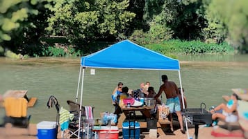Un gruppo di persone riunite attorno a un tavolo sotto una tenda blu, vicino a un fiume, con attrezzatura da campeggio e un barbecue.