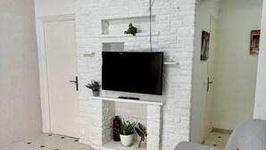 Ett vardagsrum med en vit tegelkamin, en platt-TV monterad ovanför, en grå soffa med en orange kudde och ett klinkergolv med stjärnmönster.