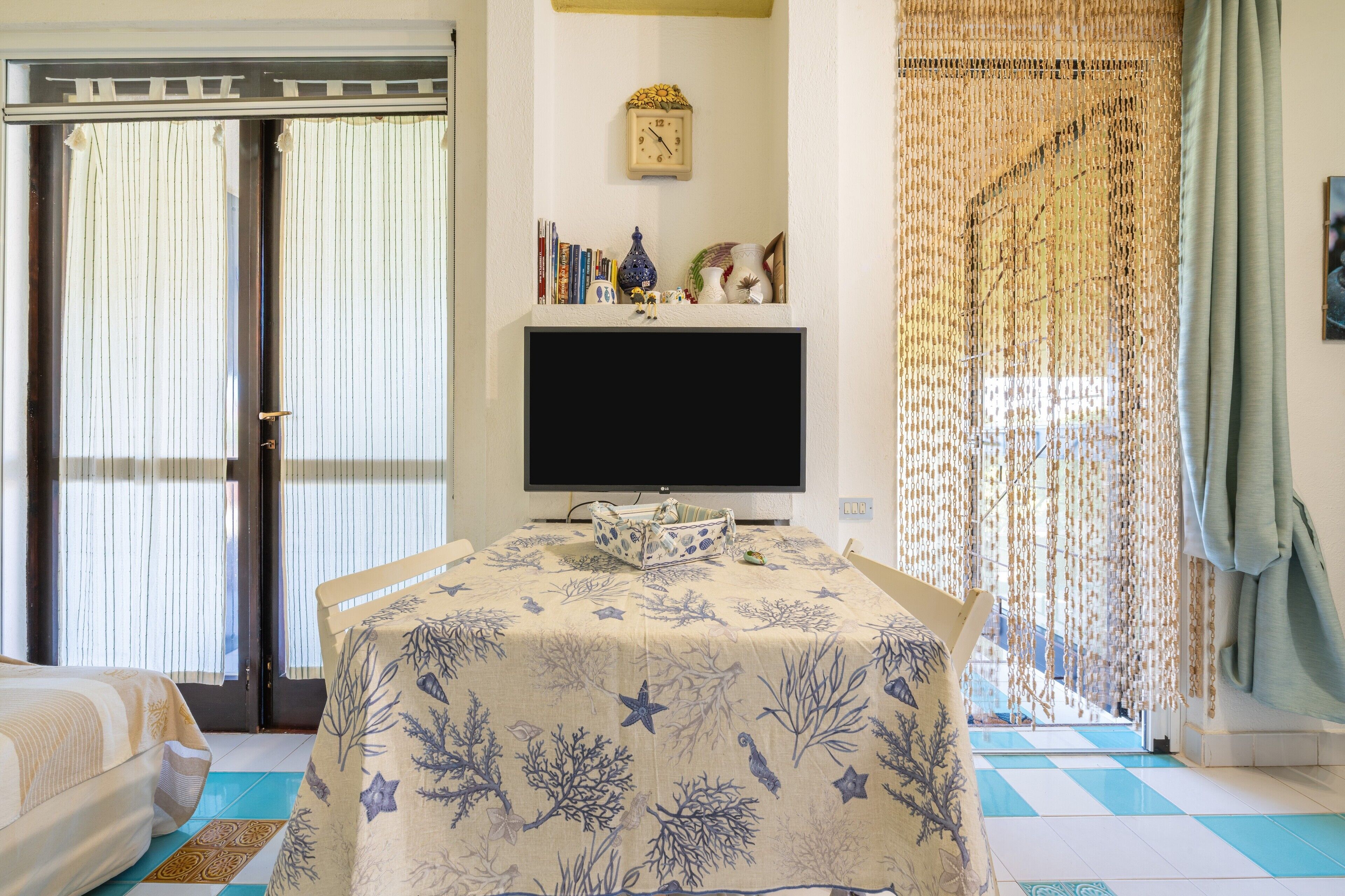 A room with a bed, a TV on a stand, a table with a tablecloth, and a window with curtains.