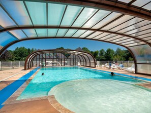 A covered swimming pool with clear blue water, a curved roof, and a view of a building in the background. - Normandy Coastal Stay for 4 (Parentis-en-Born)