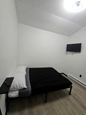 Een kleine kamer met een enkel bed, een bureau, een wandgemonteerde flatscreen-tv en een plafondlamp.