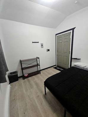 Een slaapkamer met een bed, een plank, een deur en een prullenbak.