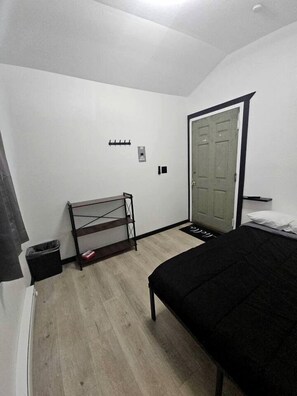 Een slaapkamer met een bed, een plank, een deur en een prullenbak.