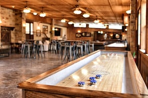 A bar area with a shuffleboard table, wooden bar stools, and hanging lights. - Reconnect by the Water: Tented Cabin Oasis for a Texas Lakeside Getaway (Cibolo)