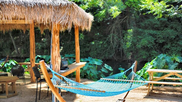 A hammock, wooden chairs, a table, and a thatched-roof structure in a garden setting. - Reconnect by the Water: Tented Cabin Oasis for a Texas Lakeside Getaway (Cibolo)