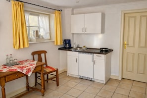 A compact kitchen with white cabinets, a black countertop, a single chair, a table with a floral tablecloth, and a window with yellow curtains. - Charming Apartments in a Half-Timbered House (Egestorf)