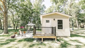 A small, single-story cabin with a front porch, surrounded by trees and a patio area with outdoor furniture.