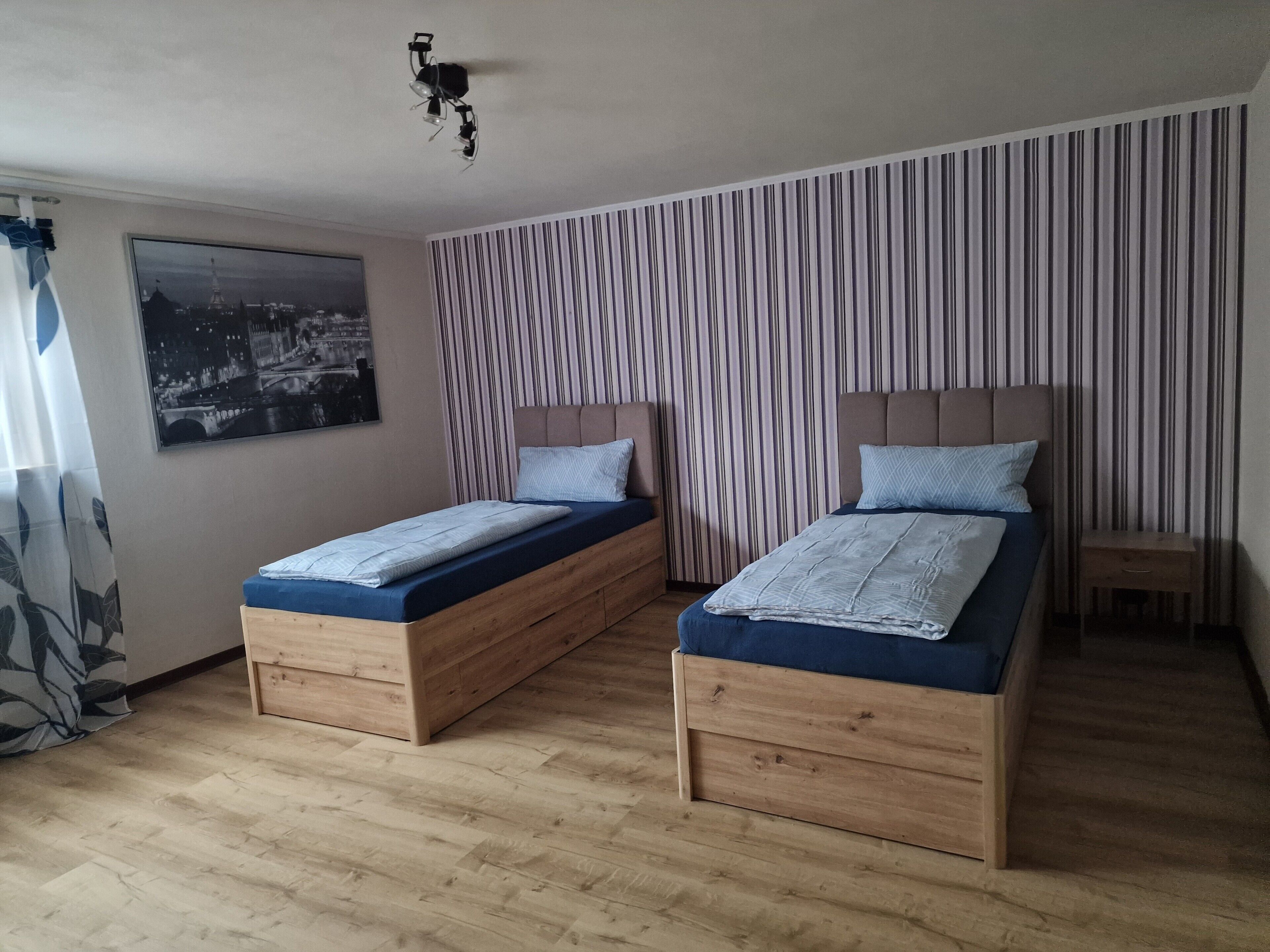 A room with two wooden beds, a striped curtain, a ceiling fan, and a cityscape painting.