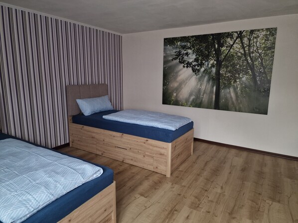 A bedroom with two beds, striped wallpaper, a forest-themed wall art, and wooden flooring.