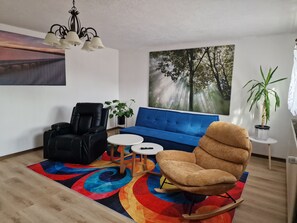 A living room with a blue sofa, black leather chair, and a colorful rug. - Vacation Room with Private Kitchen, Bathroom, and Washing Machine (Sibbesse)