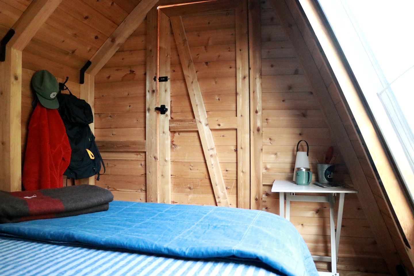 A small attic room with a sloped ceiling, a bed with a blue blanket, a ladder, and a small table with a cup and a mug.