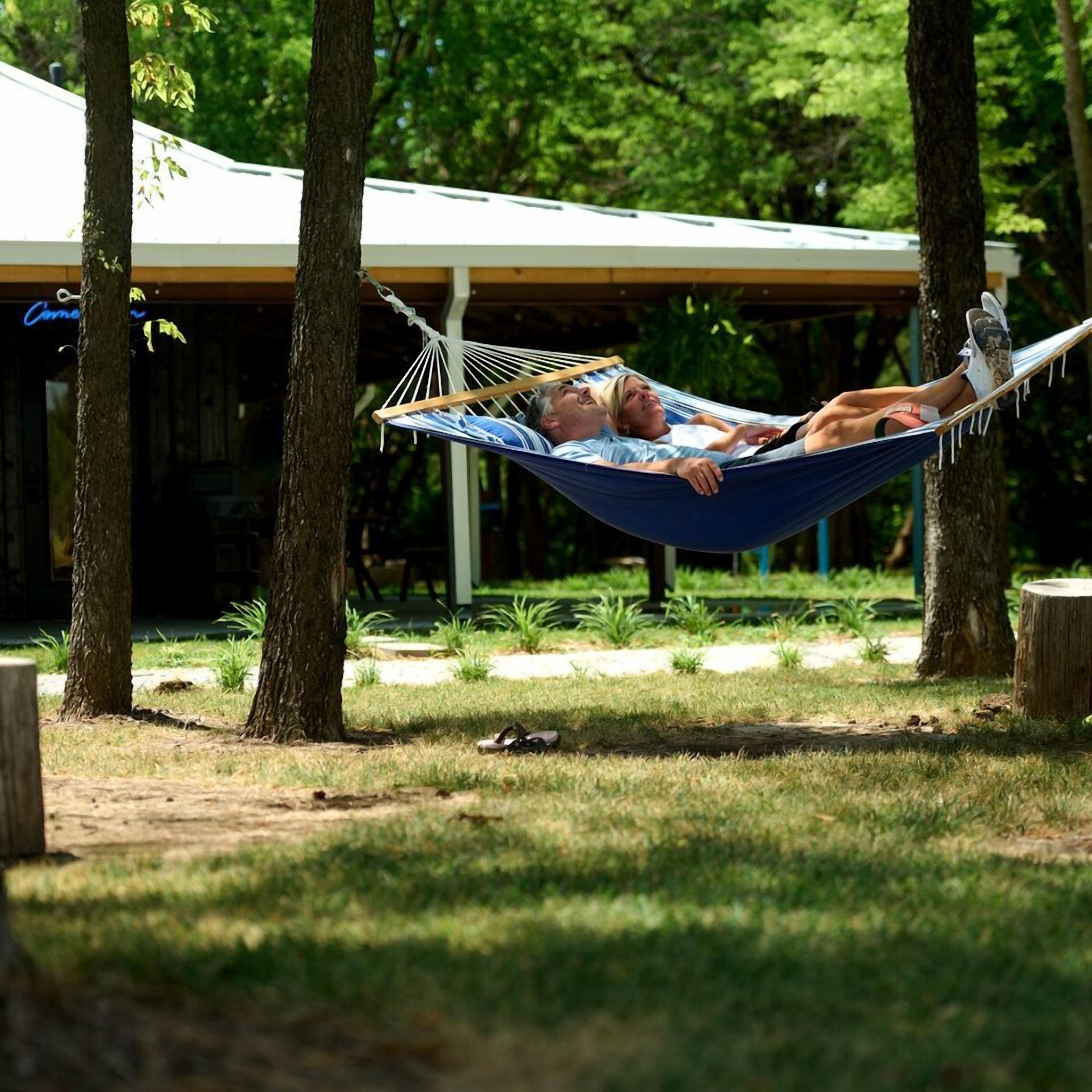 A hammock between trees in a park with a covered area in the background.