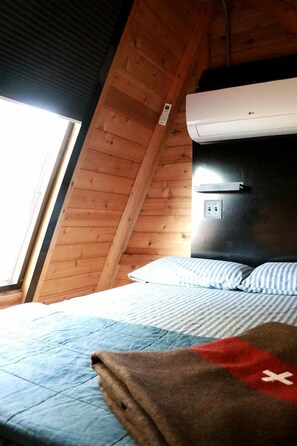A bedroom with a sloped ceiling, a bed with blue bedding, a red blanket, and an air conditioning unit mounted on the wall.