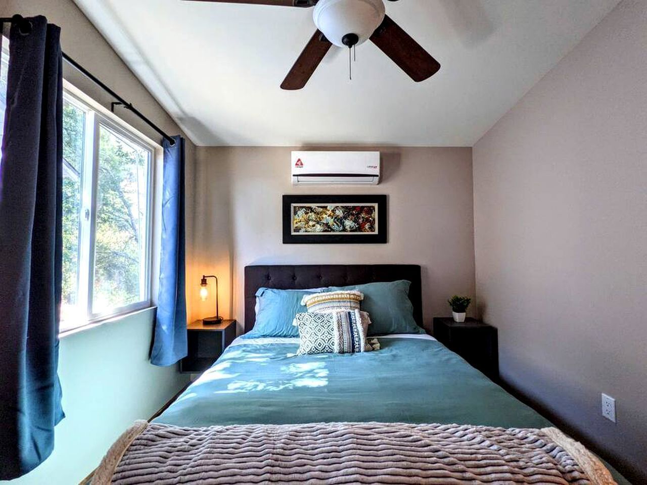 A bedroom with a bed, a ceiling fan, an air conditioner, a nightstand, a lamp, and a window with curtains.