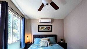 Quarto com cama, ventoinha no teto, ar condicionado, mesa de cabeceira, abajur e janela com cortinas.