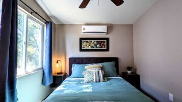 A bedroom with a bed, a ceiling fan, an air conditioner, a nightstand, a lamp, and a window with curtains.