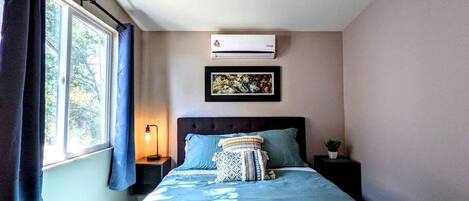 Une chambre à coucher comprenant un lit, un ventilateur de plafond, un climatiseur, une table de chevet, une lampe et une fenêtre avec des rideaux.
