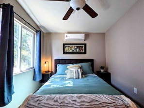 Een slaapkamer met een bed, een plafondventilator, een airconditioner, een nachtkastje, een lamp en een raam met gordijnen.