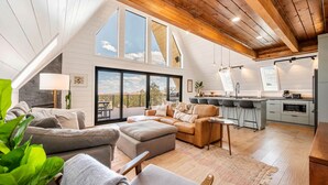 A spacious loft with a high ceiling, a cozy living area with a sofa and ottoman, a kitchen with modern appliances, and large windows offering a view of the outdoors.