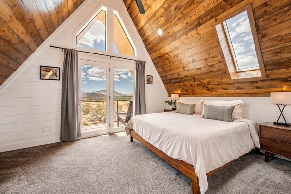 Un dormitorio con techo inclinado, una cama, una mesita de noche, una lámpara y vistas al exterior.