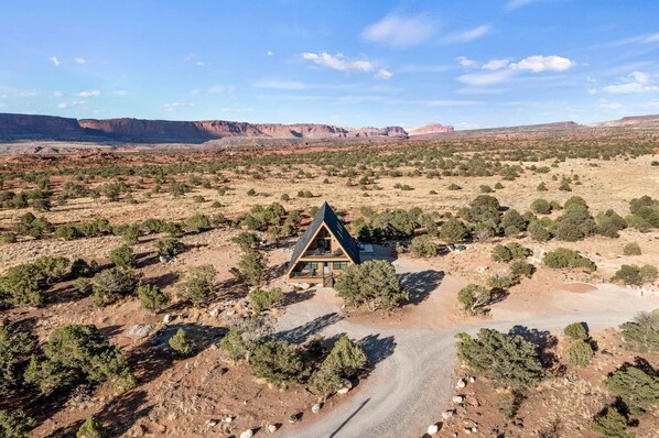 A remote cabin with a triangular roof, surrounded by sparse vegetation and mountains in the background.