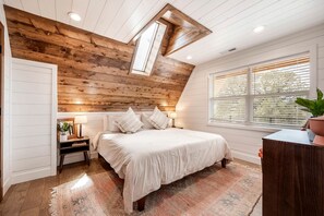 A bedroom with a sloped ceiling, a skylight, a large bed, bedside tables, a nightstand, a window with blinds, and a potted plant.