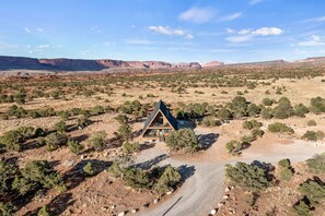 A remote cabin with a triangular roof, surrounded by sparse vegetation and mountains in the background.