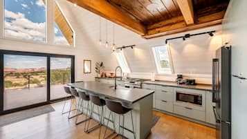 A modern kitchen with a large island, bar stools, a dining area with a view, and a high ceiling with exposed beams.