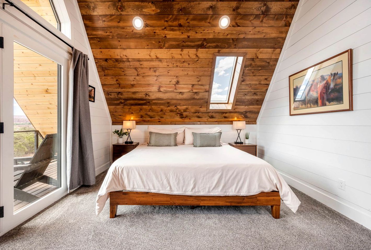 A bedroom with a wooden ceiling, a skylight, a bed with white bedding, two bedside tables with lamps, and a framed picture on the wall.