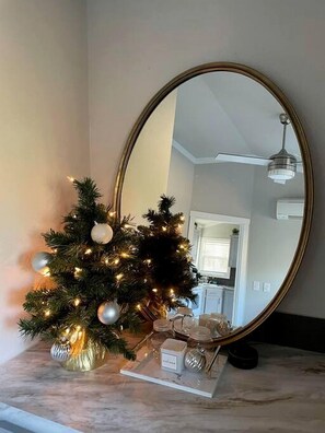 A Christmas tree with lights and ornaments on a marble countertop next to a large oval mirror.
