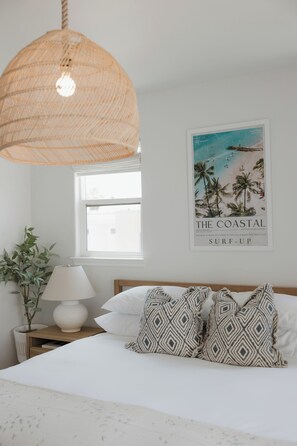 Un dormitorio con cama, lámpara colgante, un cartel en la pared, planta en la mesita de noche y una ventana.