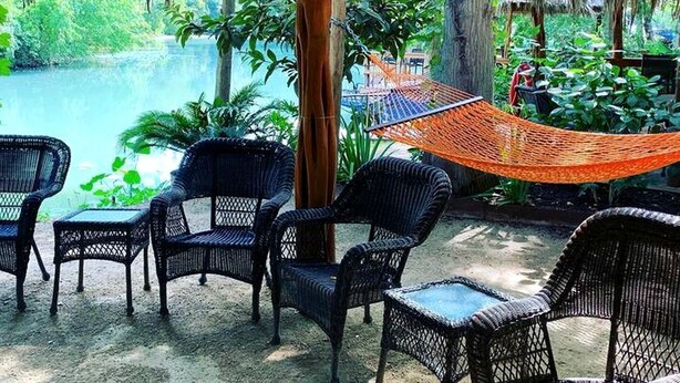 An outdoor seating area with wicker chairs and a hammock, overlooking a body of water and lush greenery. - Luxurious Safari Tent, Amazing for Glamping Vacations near Seguin, Texas (Seguin)