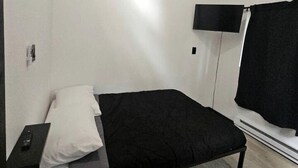 A single bed with a black mattress and white pillow, a bedside table with a remote control, a wall-mounted TV, and a window with a black curtain. - Peaceful Cozy Cabin Stay With Play Area Close To River For Fishing And Easy Outdoor Fun in Houston, Alaska (Houston)