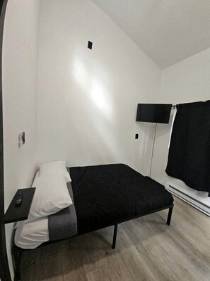A single bed with a black mattress and white pillow, a bedside table with a remote control, a wall-mounted TV, and a window with a black curtain. - Peaceful Cozy Cabin Stay With Play Area Close To River For Fishing And Easy Outdoor Fun in Houston, Alaska (Houston)