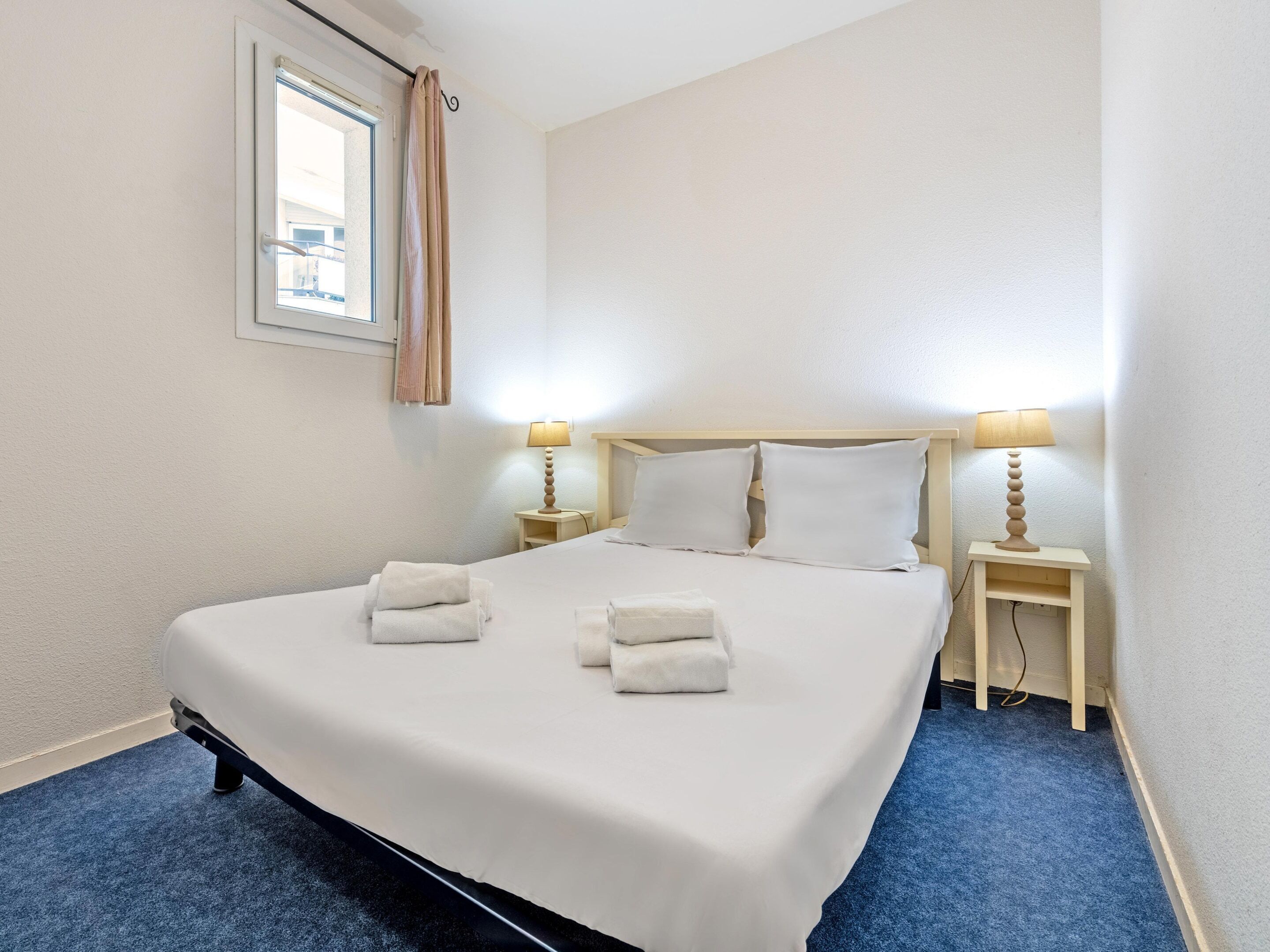 Habitación de hotel con una cama grande, dos mesitas de noche con lámparas, una ventana con cortinas y una alfombra azul.