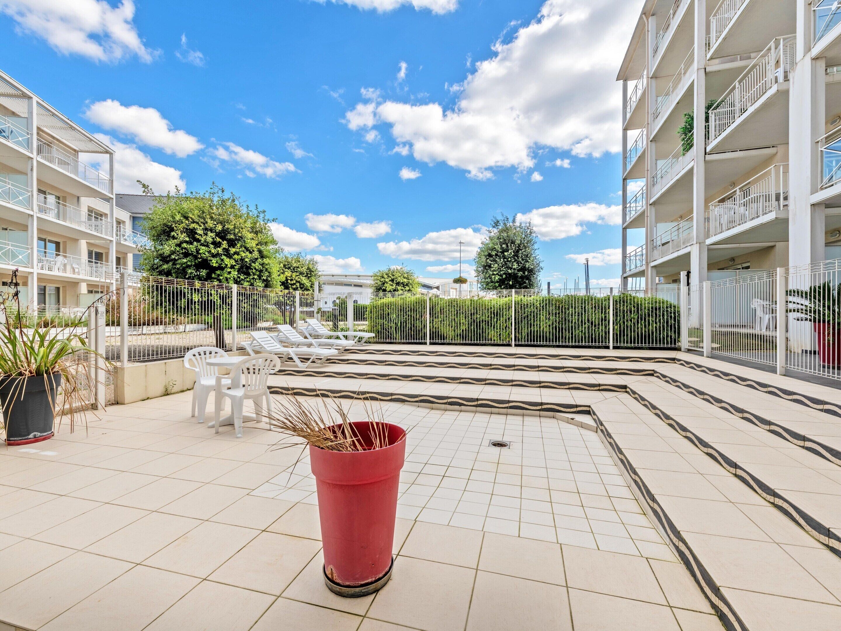 Un patio con suelo de baldosas, una maceta roja, sillones blancos y una valla de metal.