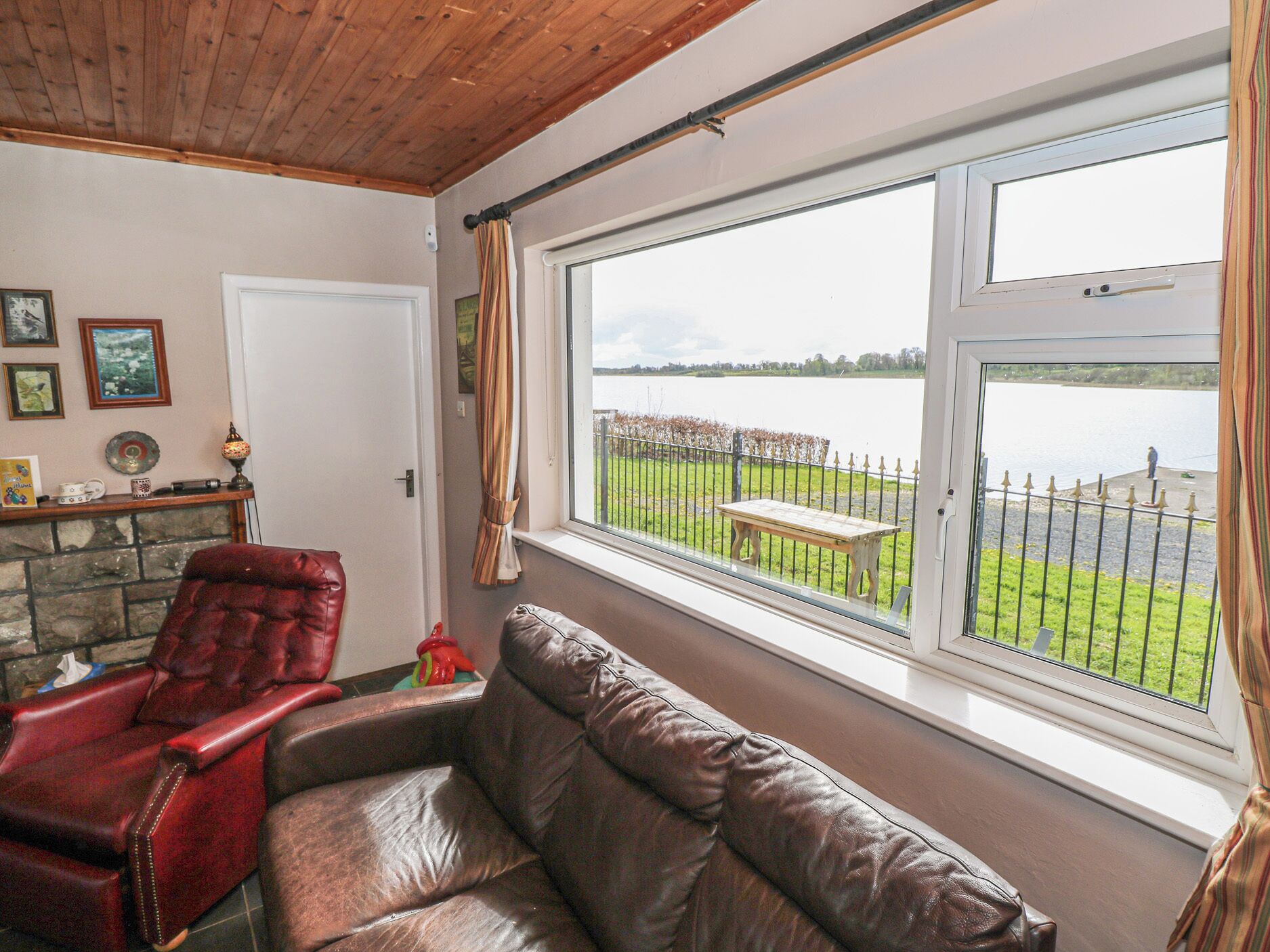 A room with a leather sofa, a red recliner, a stone fireplace, a window with a view of a lake and a bench, and a wooden ceiling.
