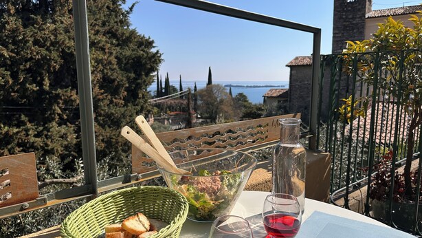 A table with a basket of bread, two glasses of red wine, and a water bottle on a balcony overlooking a scenic view of a body of water and buildings. - Lake view & tranquillity, Villa Laguna Blue, whirlpool & sauna-10 minutes walk to the lake (Gardone Riviera)