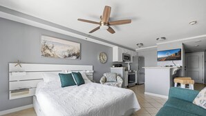 Una habitación de hotel moderna con una cama grande, ventilador de techo, televisión, kitchenette y una pequeña zona de estar.