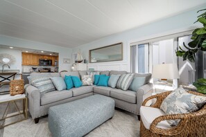 Ein Wohnzimmer mit grauem Ecksofa, Hocker und Korbstühlen. Im Hintergrund ist eine Küche mit Holzschränken und einer Mikrowelle zu sehen.