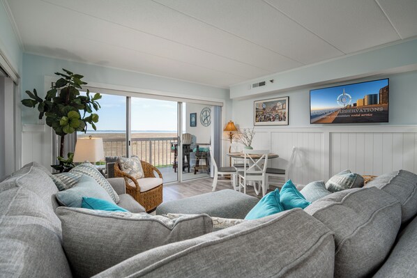 Ein geräumiges Wohnzimmer mit einer grauen Sofagarnitur, einem Korbstuhl und einem Esstisch mit Stühlen. Ein großes Fenster bietet Blick auf den Strand.