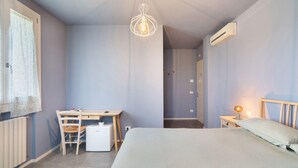 A bedroom with a bed, a desk, a chair, a hanging light fixture, and an air conditioning unit. - Melloni Street Home - Lavender Room (Dolo)