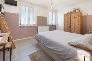 Un dormitorio con cama, armario de madera, escritorio con silla, radiador y televisor en la pared.