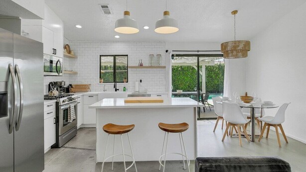 A modern kitchen with white cabinetry, stainless steel appliances, a central island with stools, and a dining area with a table and chairs. - Desert Oasis w/ Pool & Spa, Game Room + Theater (Indio)